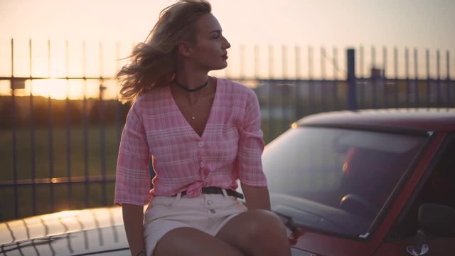 beautiful blonde young woman sits on the hood of her car at sunset