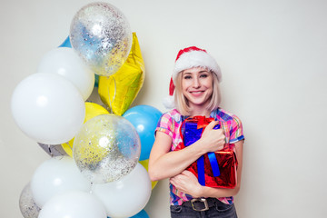 Fototapeta na wymiar portrait of young Beautyful blondу amazed woman with christmas box gift on red background. lady in a plaid shirt and santa claus hold a bag full of presents near helium multi-colored balloons studio