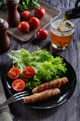 Appetizing and colorful sausages wrapped in bacon and grilled next to tomatoes and lettuce are at black ceramic plate on a wooden table next to glass of beer