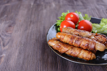 Appetizing and colorful sausages wrapped in bacon and grilled next to tomatoes and lettuce are on a wooden table