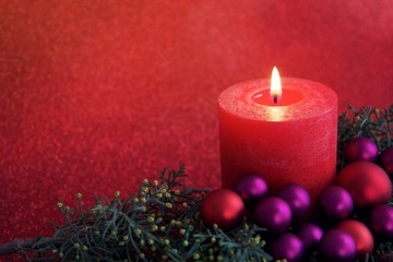 Eine rote, brennende Kerze mit Zypressenzweig und Weihanchtskugeln vor rot leuchtendem Hintergrund.