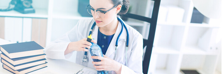 A beautiful young girl in a white robe sits at a computer desk, holds a marker in her hand and opens a bottle of water.