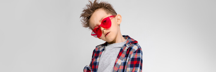 A handsome boy in a plaid shirt, gray shirt and jeans stands on a gray background. A boy in red sunglasses. The boy laid his hand on his arm.