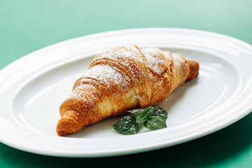One fresh croissants on white plate with powdered sugar and mint leaves on modern green background. Horizontal close view