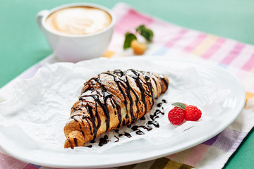 Fresh homemade croissants and cappuccino on cloth napkin decorated with raspberry and chocolate topping. Side view with copy space on green background