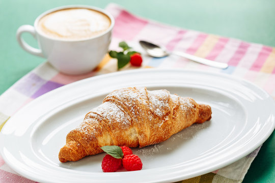 Fresh homemade croissants and cappuccino on cloth napkin decorated with raspberry and chocolate topping. Side view with copy space on green background
