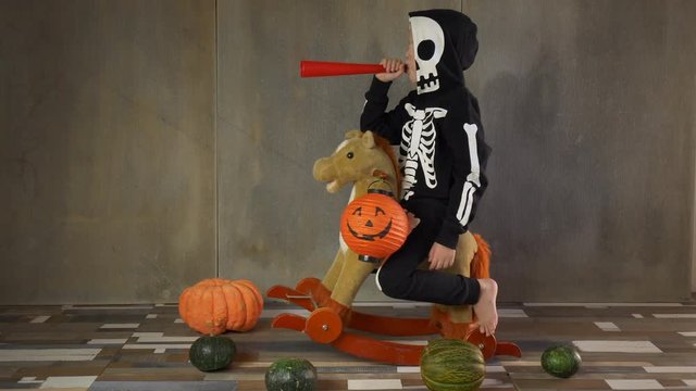 Little boy barefoot in skeleton suit on rocking toy horse. Child happily celebrates Halloween, holds pumpkin-shaped flashlight and hits red tube