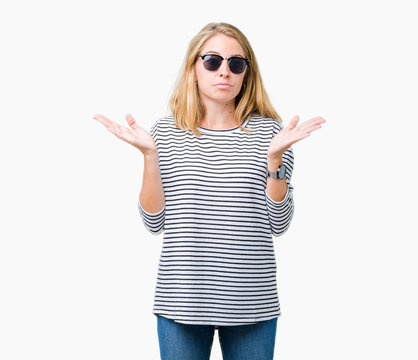 Beautiful young woman wearing sunglasses over isolated background clueless and confused expression with arms and hands raised. Doubt concept.