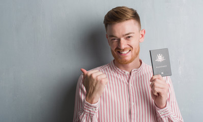 Young redhead man over grey grunge wall holding passport of Australia pointing and showing with thumb up to the side with happy face smiling