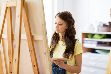 art school, creativity and people concept - student girl or young woman artist with easel and palette painting at studio