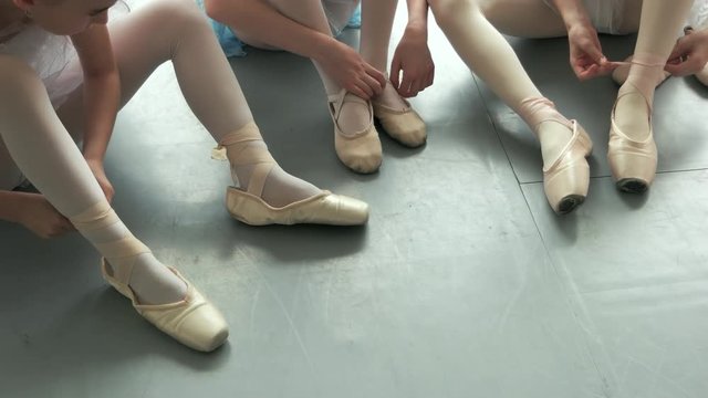 Close up little ballerinas tying their pointe shoes. Young ballerinas putting on pointe shoes while sitting on floor in ballet class.