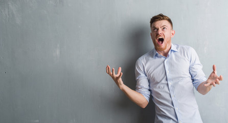 Young redhead business man over grey grunge wall crazy and mad shouting and yelling with aggressive expression and arms raised. Frustration concept.