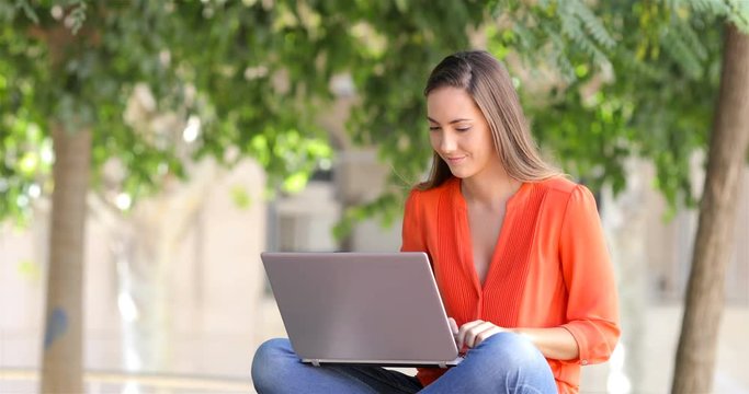 Excited woman in orange using a laptop sitting on a on a bench in a park