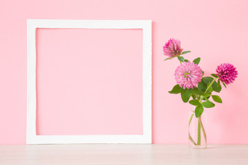 Fresh, pink clover in vase at wall. Wild flowers. Soft pastel color. Mockup for positive idea. Empty place for inspirational, emotional, sentimental text, quote or sayings in white frame. Front view.