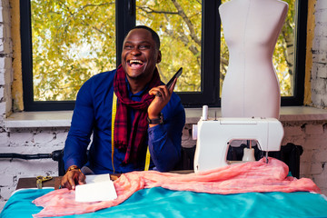 portrait of a handsome african man smiling seamstress with sewing machine.Afrio American man stylish designer working in tailor workshop mannequin,table measuring tape in room against autumn window