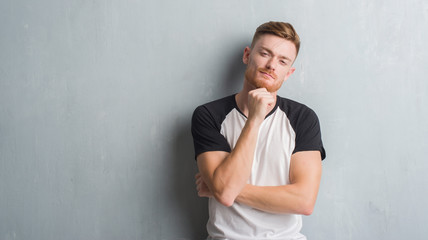 Obraz premium Young redhead man over grey grunge wall looking confident at the camera with smile with crossed arms and hand raised on chin. Thinking positive.