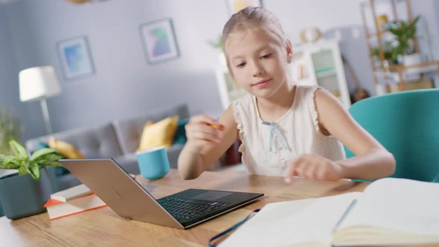 Smart Little Girl Does Homework in Her Living Room. She's Sitting at Her Desk, Uses Laptop and Writes with a Pen in Her Textbooks. 
