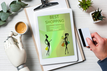 cropped image of businessman with cyborg hand holding credit card and using digital tablet with best shopping on screen at table with coffee cup in office