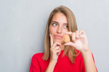 Beautiful young woman standing over grunge grey wall holding walnut serious face thinking about question, very confused idea