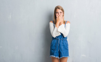 Beautiful young woman standing over grunge grey wall shocked covering mouth with hands for mistake. Secret concept.