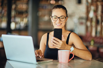 Caucasian woman using smart phone and laptop while sitting in restaurant. On desk laptop and mug with tea.
