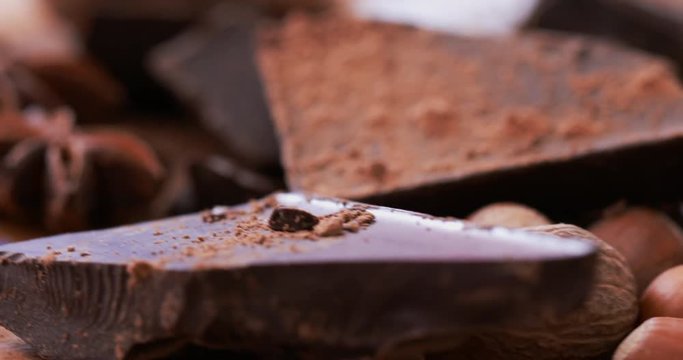 Crushed dark chocolate bar sprinkled with cocoa powder, with hazelnuts, nutmeg, walnut, chocolate candy and cinnamon at rotating stand. Extreme close up