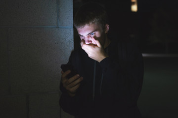 Surprised teenager covering his mouth after viewing something shocking on his phone. He is leaning against the brick wall of a high school at night.