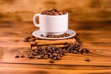 White cup filled with coffee beans, star anise and cinnamon sticks on wooden table