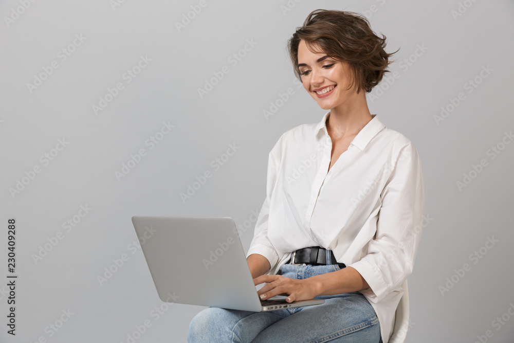 Wall mural Young business woman posing isolated over grey wall background sitting on stool using laptop computer.