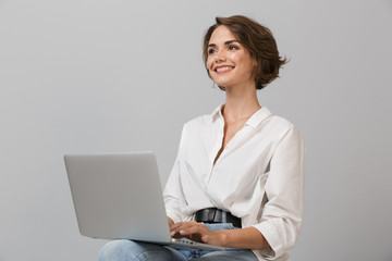 Young business woman posing isolated over grey wall background sitting on stool using laptop...