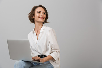 Young business woman posing isolated over grey wall background sitting on stool using laptop...