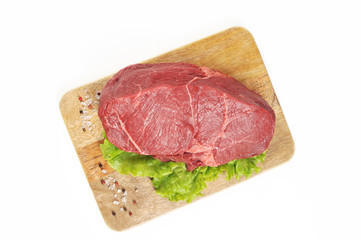 A slice of raw beef meat on a wooden board. The meat is decorated with a leaf of green salad and spices. White background. Close-up. View from above.