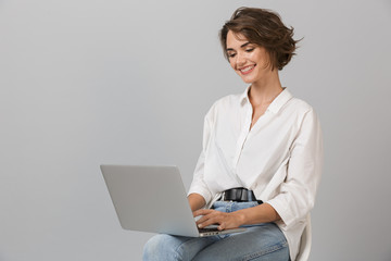 Young business woman posing isolated over grey wall background sitting on stool using laptop...