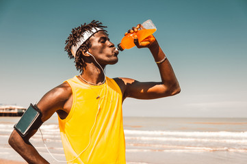 Black runner taking rest drinking energy drink at the beach at summer - obrazy, fototapety, plakaty