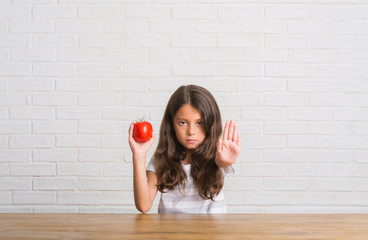 Young hispanic kid sitting on the table eating fresh tomato with open hand doing stop sign with...