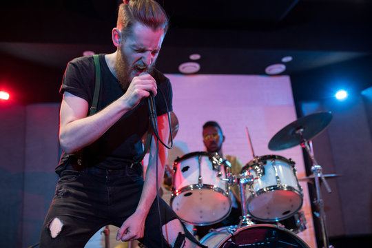 Portrait of contemporary rock musician singing fiercely on stage with drummer in background, copy space