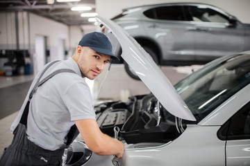 Handsome young worker looks on camera. He makes repairs in front body of car. Guy wears cap and uniform.