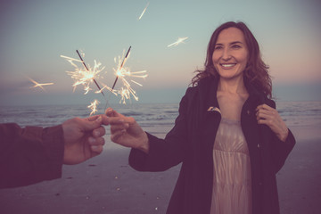 Young happy smiling woman holding burning Bengal light standing on sea side - two hands with sparklers (hand held fireworks) - Christmas and new year celebration and winter vacation concept