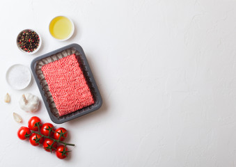 Tray with raw minced homemade beef meat with spices and herbs. Top view and space for text. On top of white kitchen table background. With pepper salt and tomatoes.