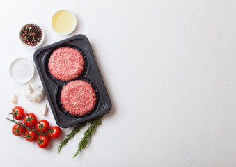 Plastic tray with raw minced homemade beef burgers with spices and herbs. Top view and space for text on top of white kitchen table background with tomatoes salt and pepper.
