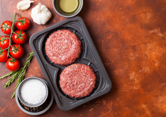 Plastic tray with raw minced homemade beef burgers with spices and herbs. Top view and space for text on rusty kitchen table background with tomatoes salt and pepper.