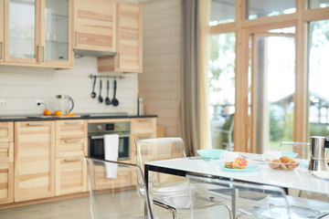 Empty domestic kitchen with modern table setting for breakfast and transparent chairs, wooden kitchen set in background
