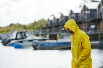Serious sad young fisherman in yellow waterproof coat holding hands in pockets and looking down...