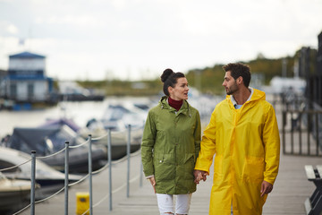 Content beautiful young couple in waterproof coats holding hands and chatting while walking over boat dock in autumn