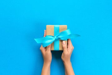 Woman arms holding gift box with blue ribbon on color background, top view