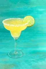 A photo of a lemon Margarita cocktail in the traditional glass with a wedge of lime on a vibrant teal background with a place for text