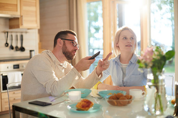 Young bearded Caucasian man pointing at TV while sitting at kitchen table with his wife, having breakfast and watching morning news attentively