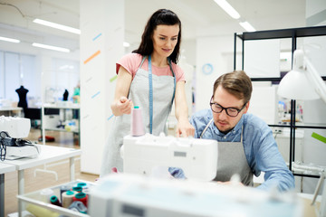 Content confident attractive young woman in apron standing at interns workplace and explaining student how to work on sewing machine in workshop