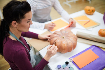 Serious concentrated female artist in apron sitting at table and painting pumpkin in art studio, she making Halloween design for food