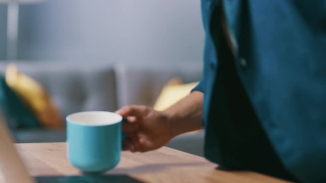 Anonymous Man Enters His Living Room with a Mug and Sits Down at His Desk Starts Working on a Laptop. 